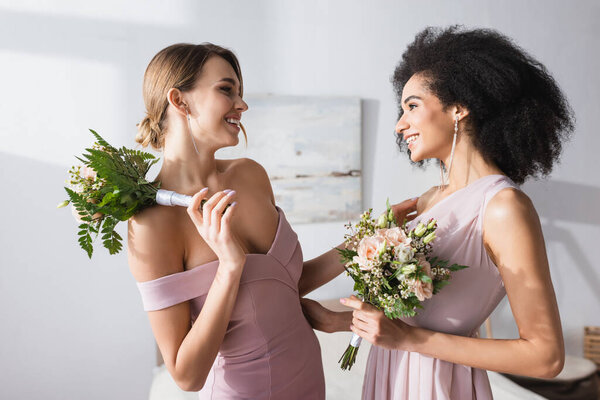 happy interracial bridesmaids smiling at each other while holding wedding bouquets