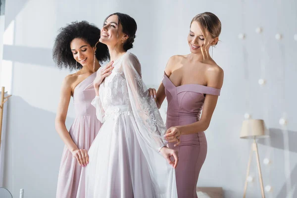 Joyful Bride Holding Hand Chest While Multicultural Bridesmaids Adjusting Her — Stock Photo, Image