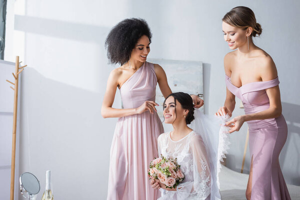 interracial bridesmaids fixing veil of happy bride sitting in bedroom with wedding bouquet 