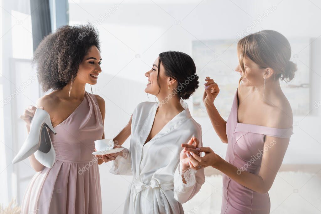 cheerful woman in silk robe smiling near interracial friends preparing her for wedding