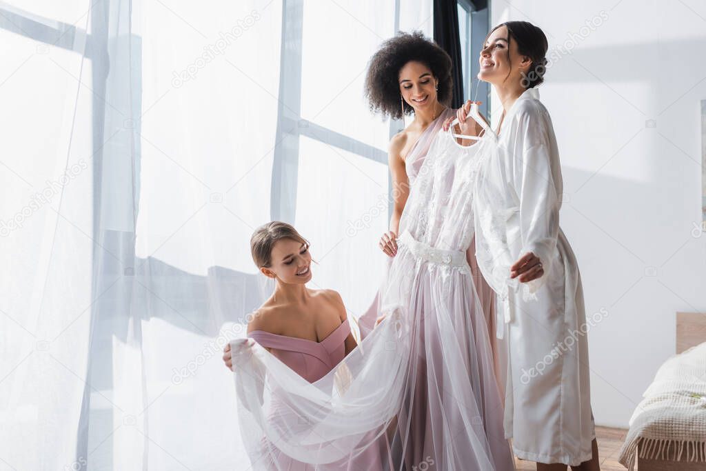 happy woman holding wedding dress near interracial bridesmaids 