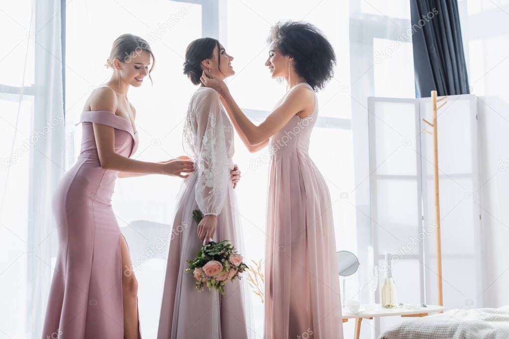 multicultural bridesmaids adjusting dress of happy bride holding wedding bouquet