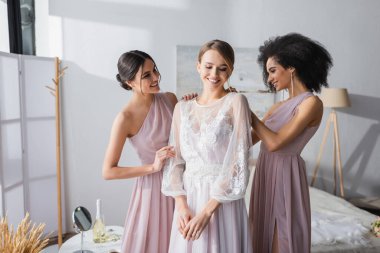 interracial bridesmaids dressing happy bride in bedroom clipart