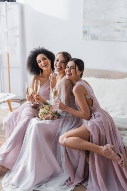 cheerful bride with bridesmaids smiling at camera while sitting with champagne in bedroom clipart