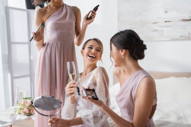 excited bride laughing near interracial friends holding decorative cosmetics clipart