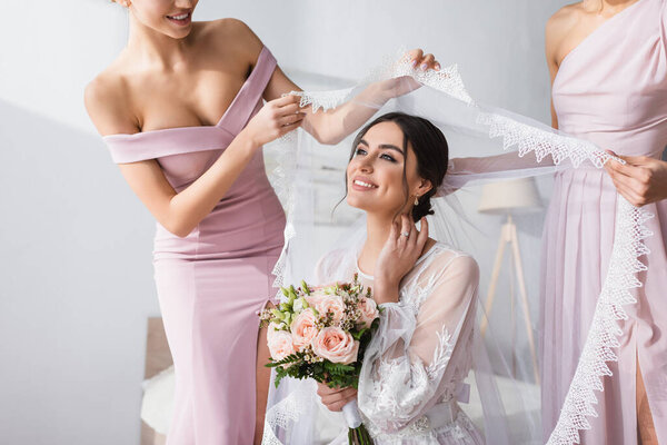 bridesmaids holding veil over pleased bride with wedding bouquet in bedroom
