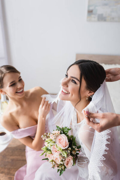 счастливая невеста, держащая свадебный букет, пока подружки невесты чинят ее вуаль