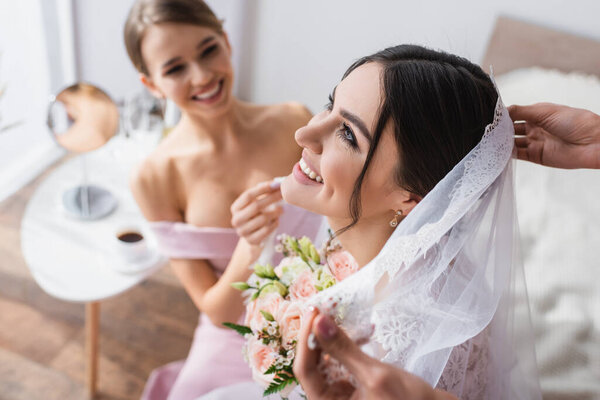 happy woman with wedding bouquet near bridesmaids fixing her veil