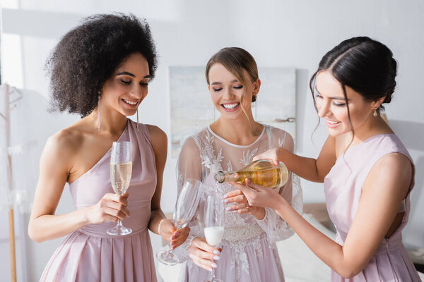 pretty woman pouring champagne near bride and african american bridesmaid