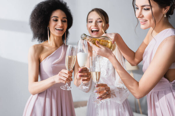 young woman pouring champagne near african american friend and bride on blurred background