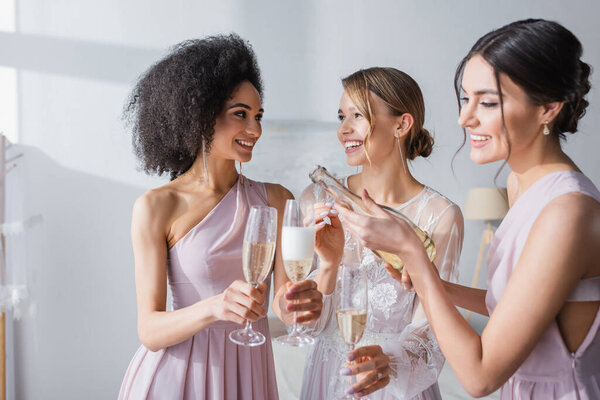 smiling woman pouring champagne near happy bride and african american bridesmaid