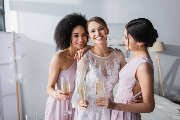 joyful bride looking at camera while holding champagne together with interracial friends
