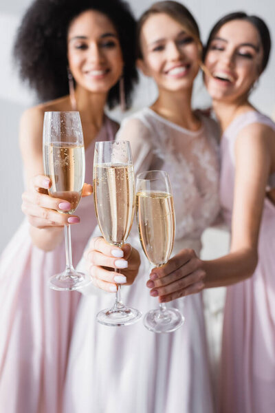 happy bride with interracial bridesmaids holding champagne glasses on blurred background