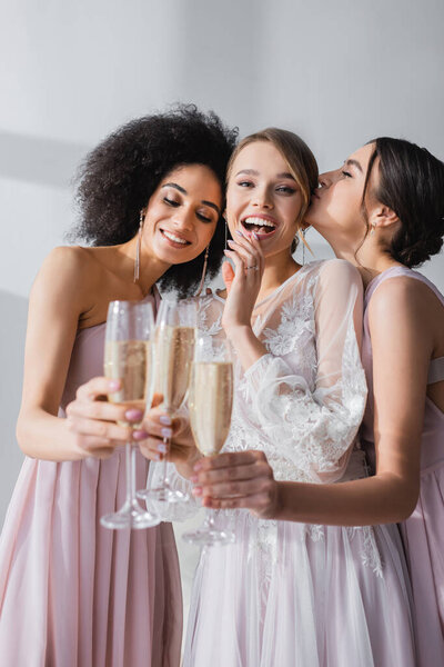 excited bride looking at camera while clinking champagne glasses with multicultural friends