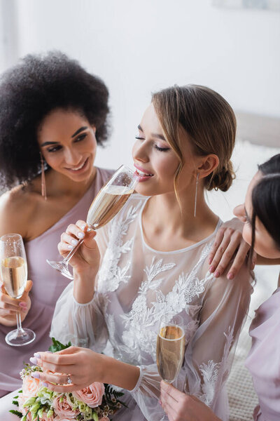 pretty bride drinking champagne near multicultural bridesmaids in bedroom