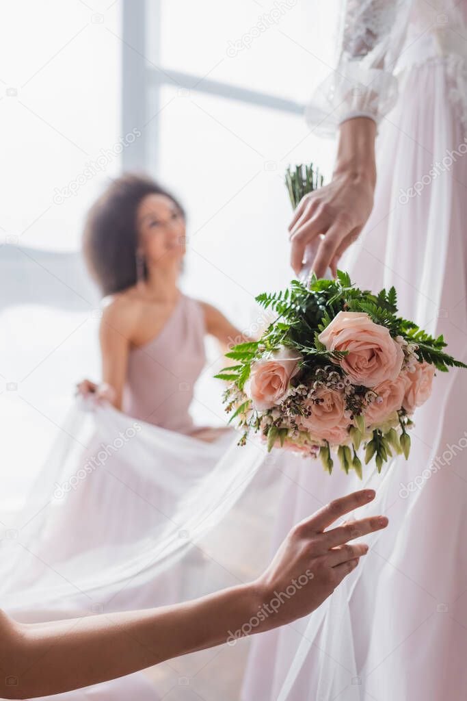 selective focus of wedding bouquet in hand of bride near african american bridesmaid on blurred background