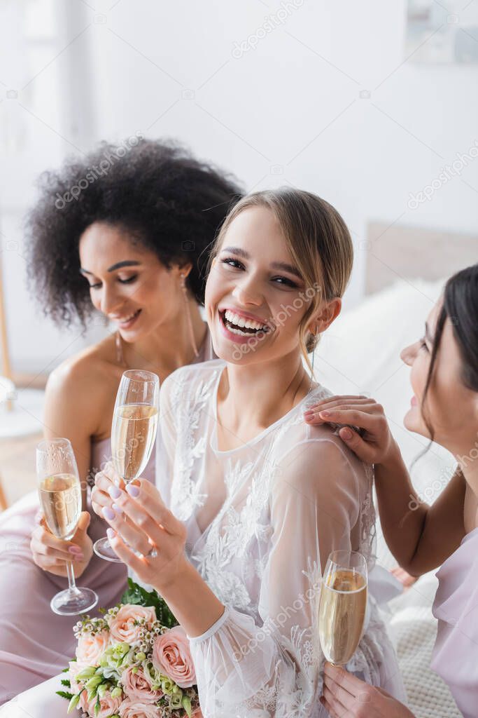laughing bride looking at camera while holding champagne near interracial friends