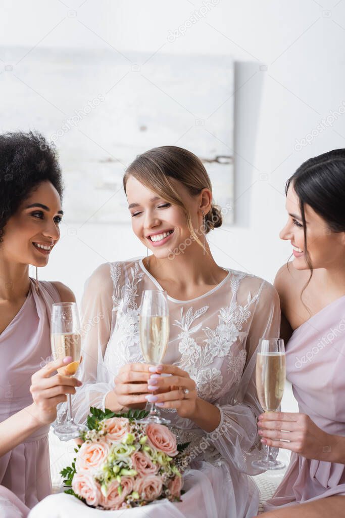 multicultural bridesmaids looking at happy bride while holding champagne glasses