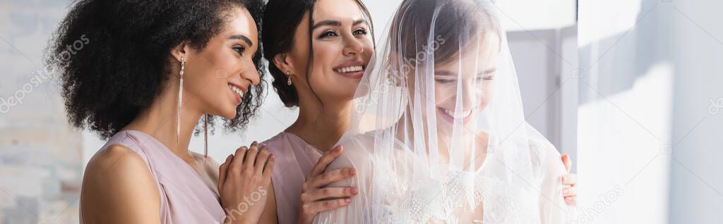 young bride in veil laughing near happy interracial bridesmaids, banner