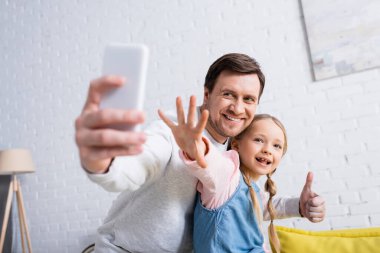 Mutlu adam, kızıyla cep telefonunda selfie çekerken baş parmağını gösteriyor, ön plan bulanık.