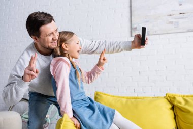 Neşeli baba ve kız akıllı telefondan selfie çekerken zafer jestleri yapıyorlar.
