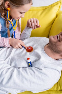 Kızının yanındaki koltukta yatan gülen adam onu oyuncak steteskopla muayene ediyor.