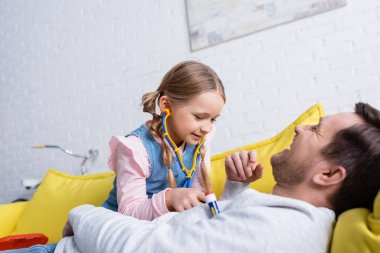Doktorculuk oynayan ve gülen babasını oyuncak steteskopla muayene eden bir kız.