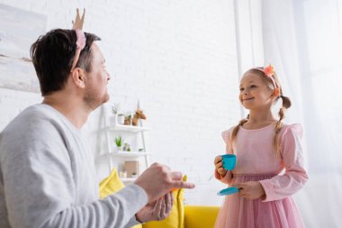 Oyuncak taclı neşeli kız bulanık önplanda babasına bakıyor.
