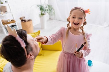 excited girl in toy crown doing makeup to dad on blurred foreground clipart