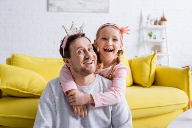 Oyuncak taçlı neşeli baba ve çocuk oturma odasında kameraya gülüyorlar.