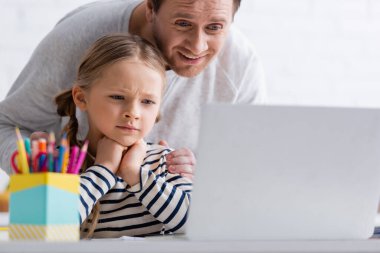 Gülümseyen bir adam, internet dersinde düşünceli kızına dokunuyor. Bulanık ön planda.