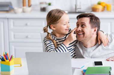 Mutfakta dizüstü bilgisayarın yanında eğlenirken babasının burnuna dokunan neşeli kız.