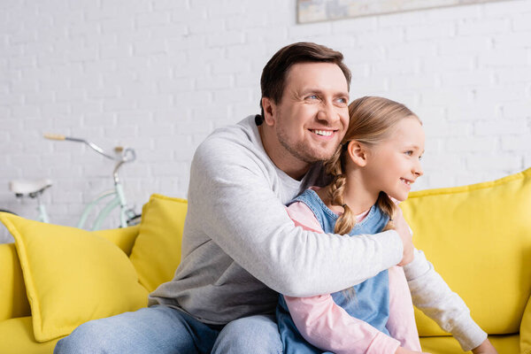 радостный мужчина смотрит в сторону, обнимая дочь на диване дома