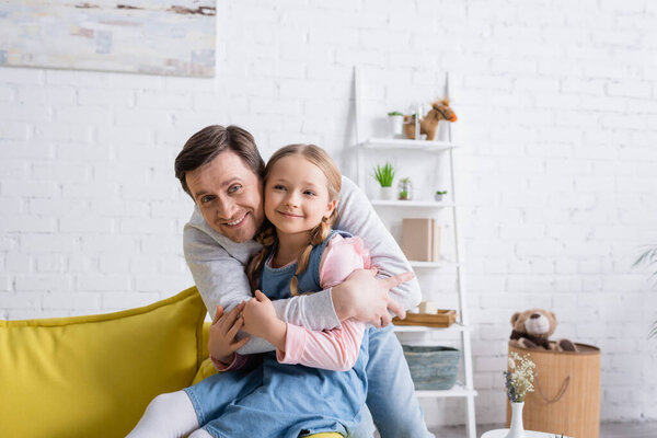 весёлый мужчина обнимает улыбающуюся дочь, сидящую дома на диване