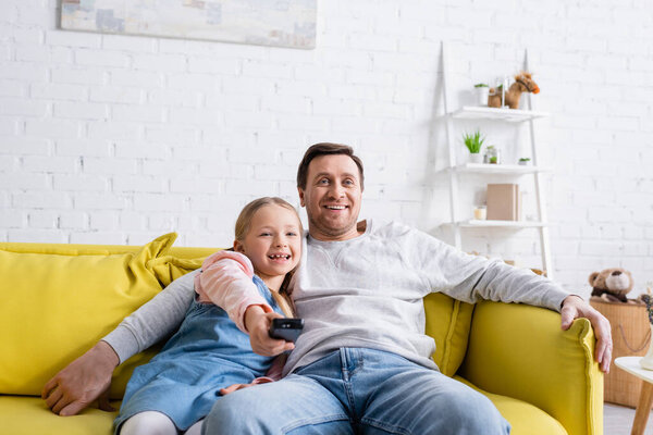 улыбающийся человек смотрит телевизор с дочерью щелкая каналы на пульте дистанционного управления
