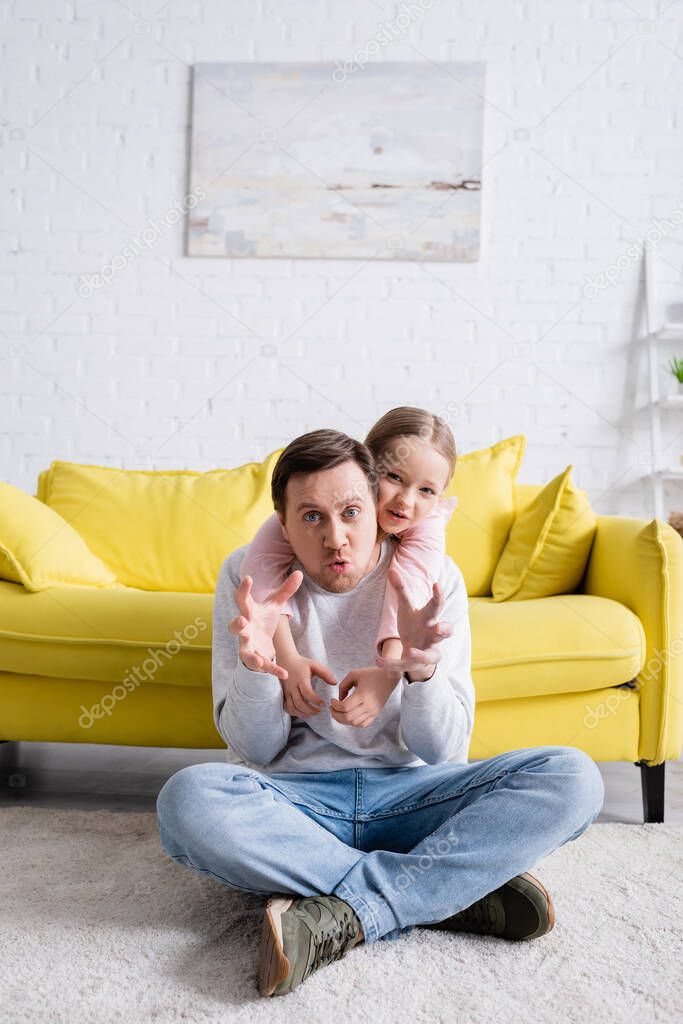 man sitting on floor and showing frightening gesture near cheerful daughter