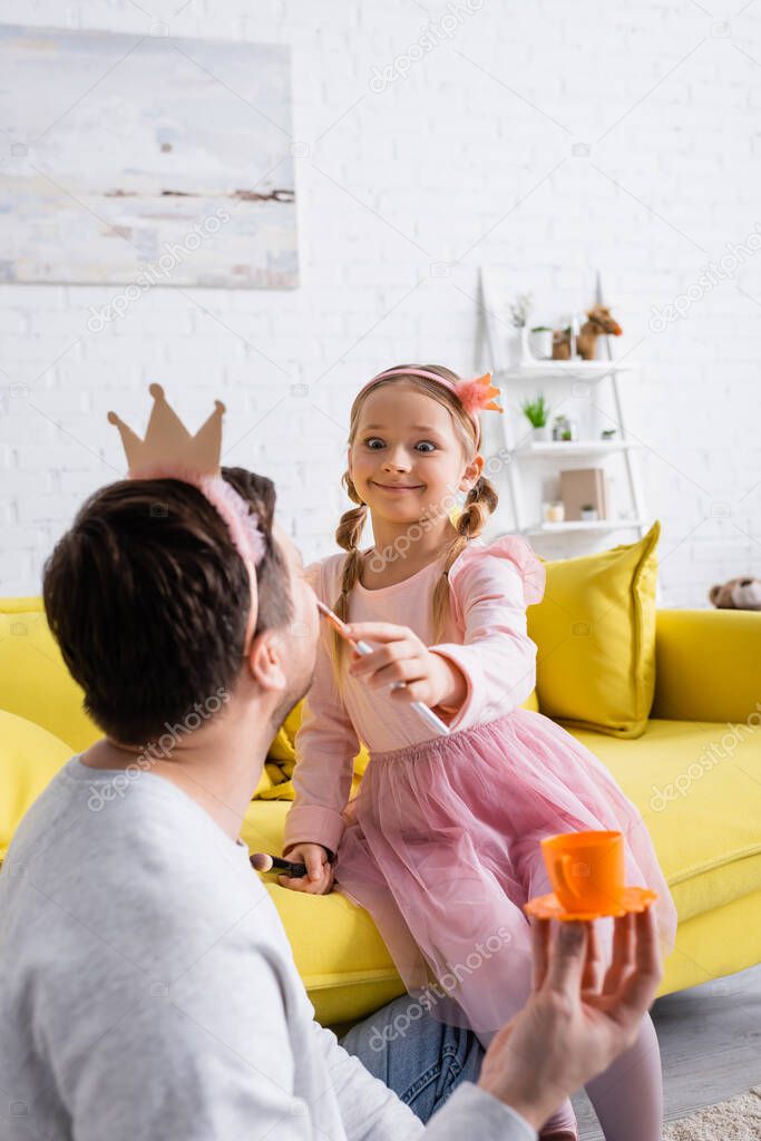 excited girl doing makeup to father holding toy cup on blurred foreground