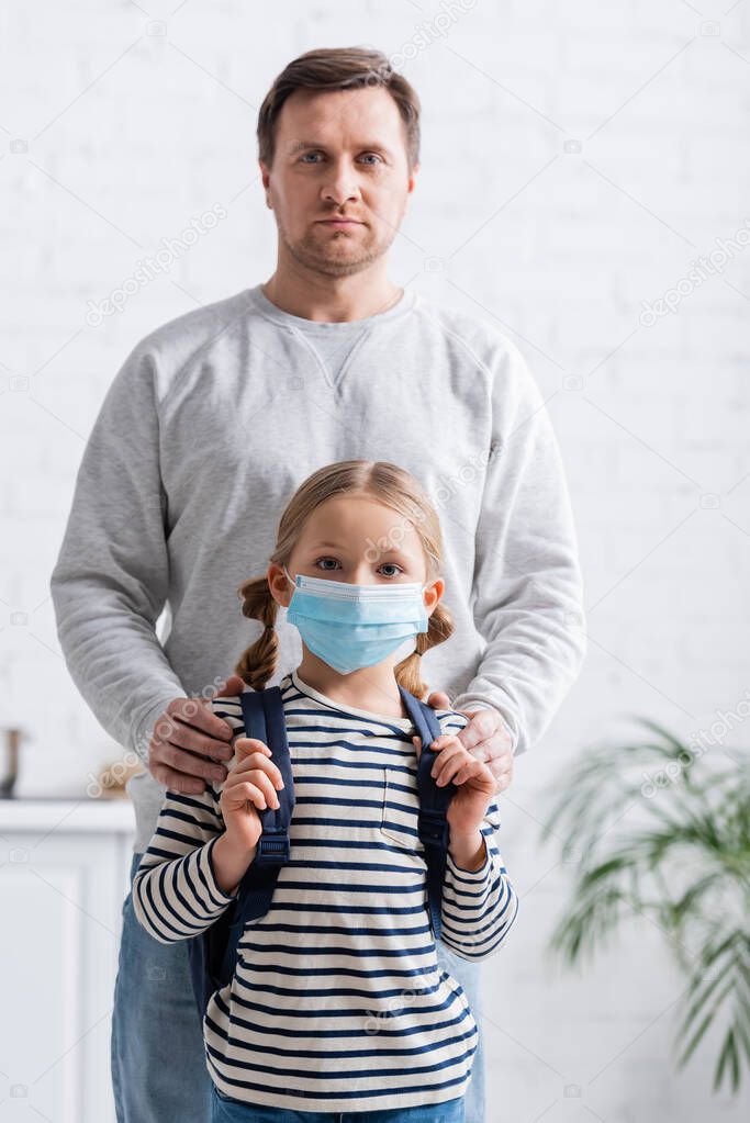 man touching shoulders of schoolgirl in medical mask while looking at camera