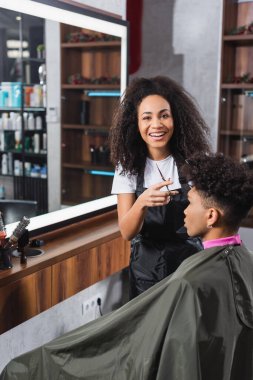 African american hairstylist with scissors smiling at camera near client in salon  clipart