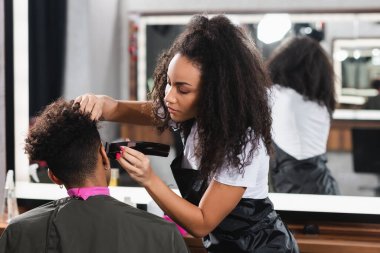 African american hairdresser in apron trimming head of client in salon  clipart