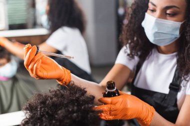 Plastik eldivenli Afro-Amerikan kuaförünün ellerinde kozmetik yağı ve bulanık ön plandaki müşteri 