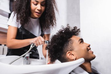 Positive african american client sitting near hairdresser with shower on blurred background in salon  clipart