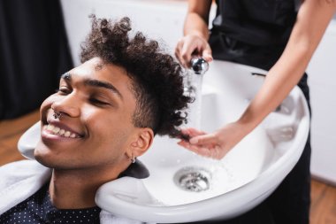 Positive african american man sitting near sink and blurred hairdresser  clipart