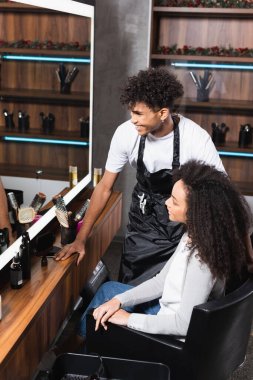 Pozitif Afro-Amerikalı kuaför müşterinin yanındaki aynaya bakıyor. 