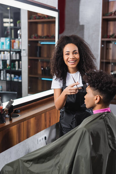 African american hairstylist with scissors smiling at camera near client in salon 