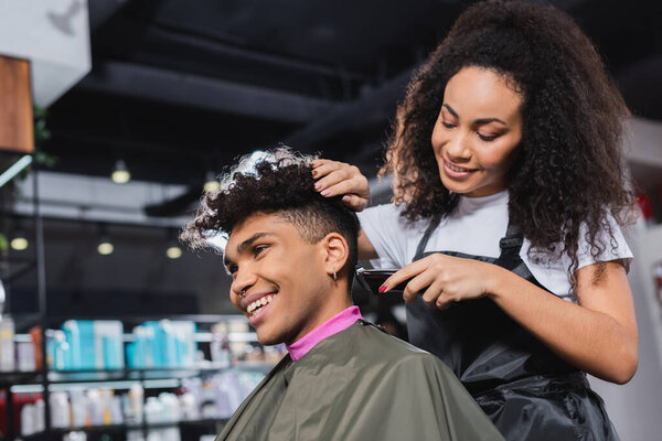 Smiling african american hairdresser in apron trimming neck of client in salon 