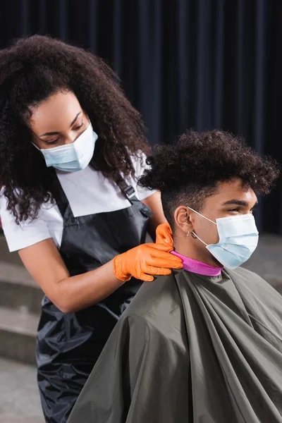 クライアントの首に襟紙を身に着けている医療用マスクでアフリカ系アメリカ人の美容師 — ストック写真