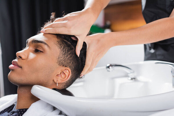 Hairstylist washing head of african american client near sink in salon 