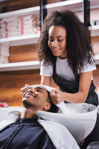 Smiling african american hairdresser holding towel near head of client in salon 