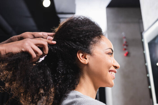 Smiling african american woman sitting near hairdresser touching curly hair 
